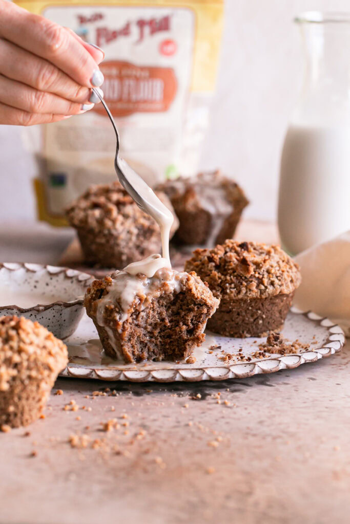 action shot of one iced cinnamon streusel muffin with a bite taken out of it, icing dripping off of a spoon and other unfrosted muffins around it, a bowl of icing beside it and a bag of Bob's Red Mill blanced almond flour in the background