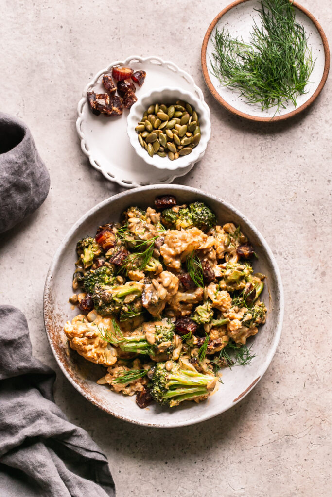 a bowl of tahini harissa cauliflower with a bowl of toasted pumpkin seeds, chopped dates, and fresh dill above it and a linen napkin on the side
