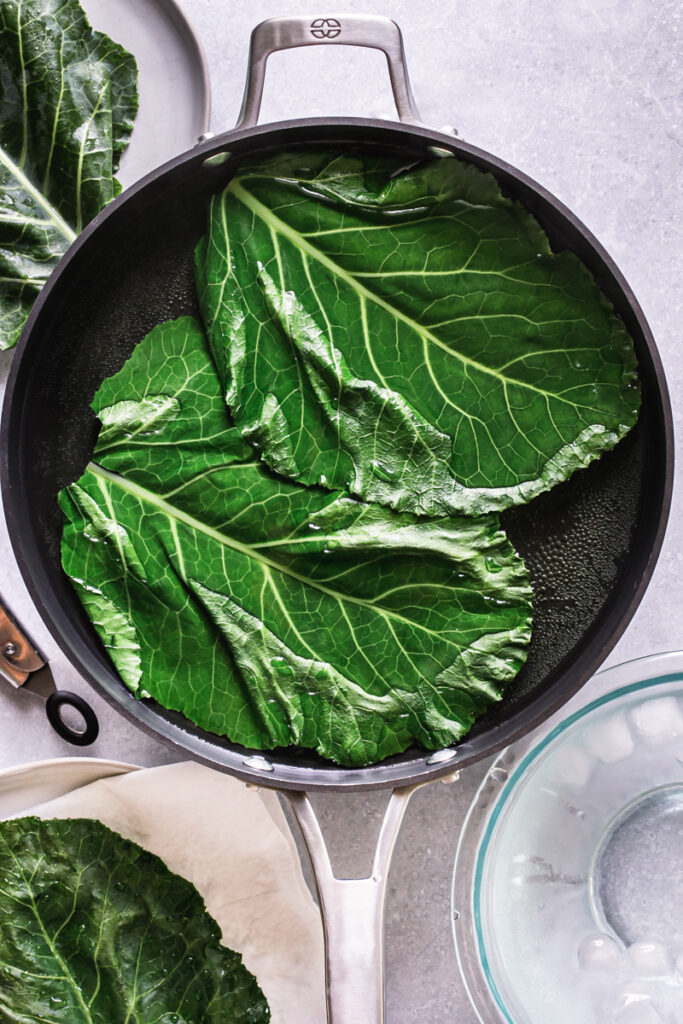 two collard wraps boiling in a skillet filled with hot water to blanch them with a bowl of ice water beside it