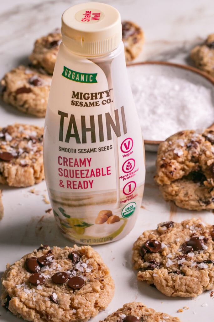 3/4 view of a bottle of mighty sesame co organic tahini with  flourless salted tahini chocolate chip cookies surrounding it and a plate of salt