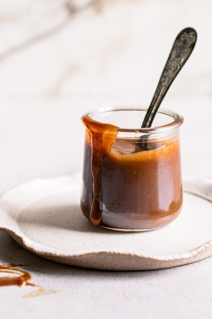 a jar of caramel on a plate with a spoon in it and the caramel dripping down the outside of the glass