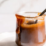 close up of a jar of caramel on a plate with a spoon in it and the caramel dripping down the outside of the glass