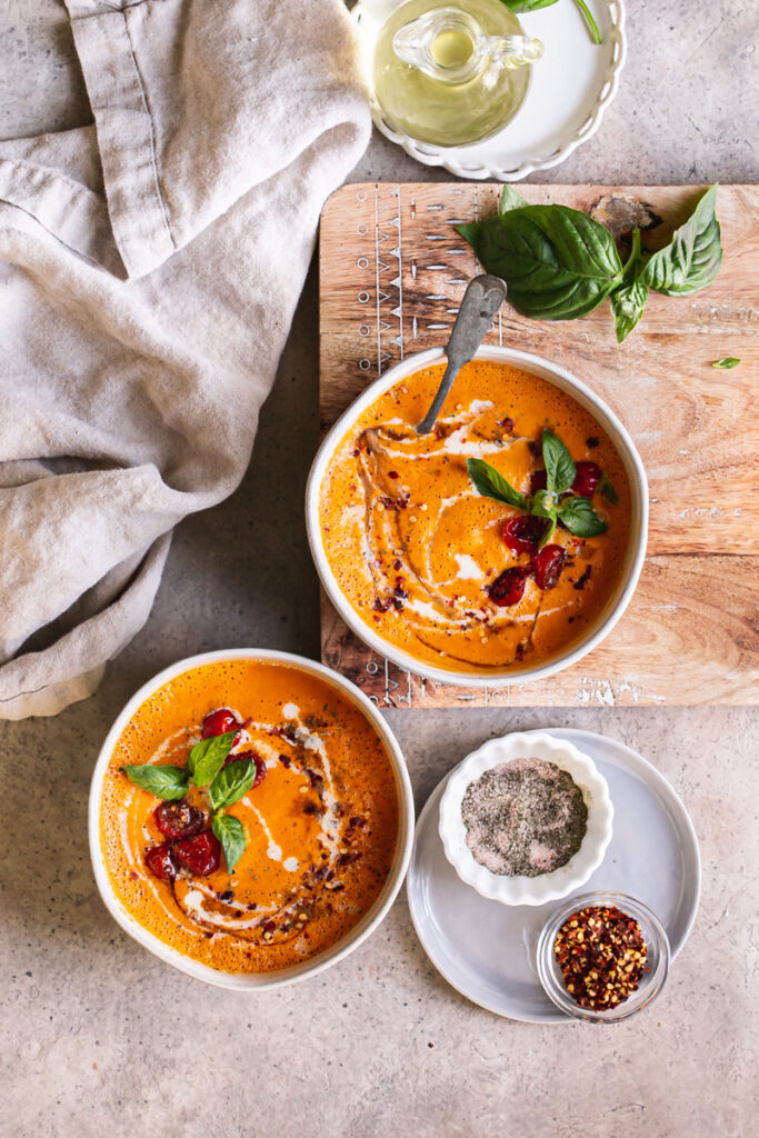 two bowls of roasted creamy tomato carrot soup with a plate of salt and pepper, red pepper flakes, and basil