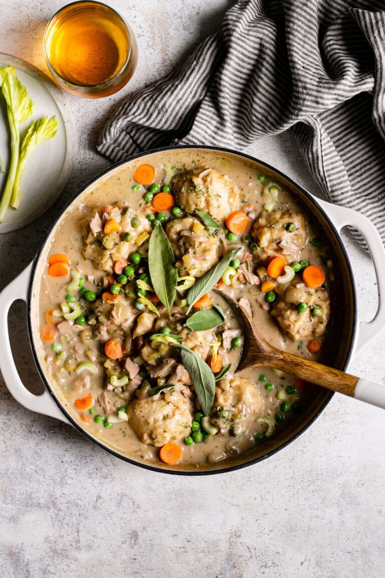 chicken and dumplings in a round Dutch Oven with a spoon in it, a glass of wine, and kitchen bowl beside it