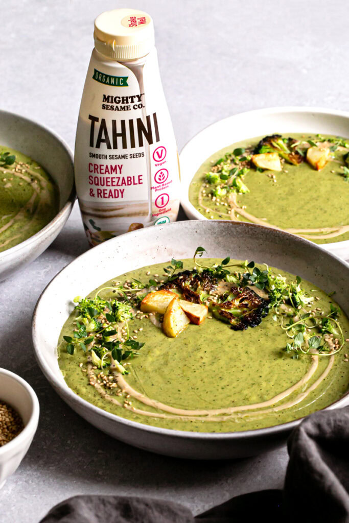 a 3/4 view of a bowl of roasted broccoli potato tahini soup with a bottle of mighty sesame co. organic tahini behind it