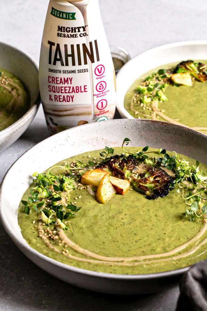 a 3/4 view of a bowl of roasted broccoli potato tahini soup with a bottle of mighty sesame co. organic tahini behind it