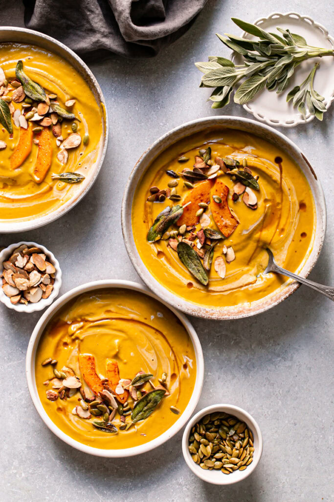 three bowls of roasted butternut squash carrot soup garnished with a plate of sage, a bowl of roasted almonds, and toasted pumpkin seeds 