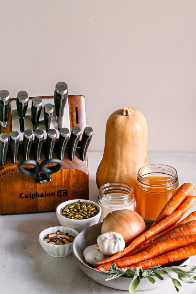 Calphalon 15-piece knife block, a whole butternut squash, a jar of vegetable broth, a jar of plant milk, a bowl of roasted almonds, a bowl of toasted pumpkin seeds, a bowl of whole carrots, an onion, garlic, and sage