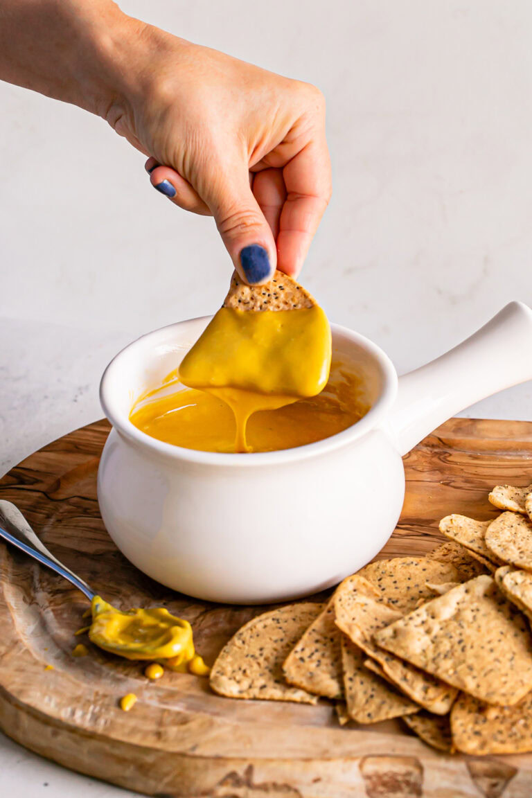 a bowl of vegan nacho cheese sauce with a chip being dipped in it and cheese sauce dripping off of the chip