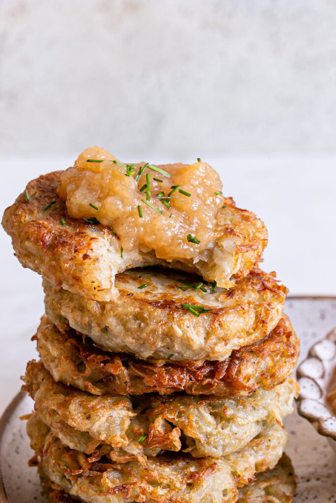 close up of potato pancakes stacked on each other, the top one topped with applesauce and a bite taken out of it.