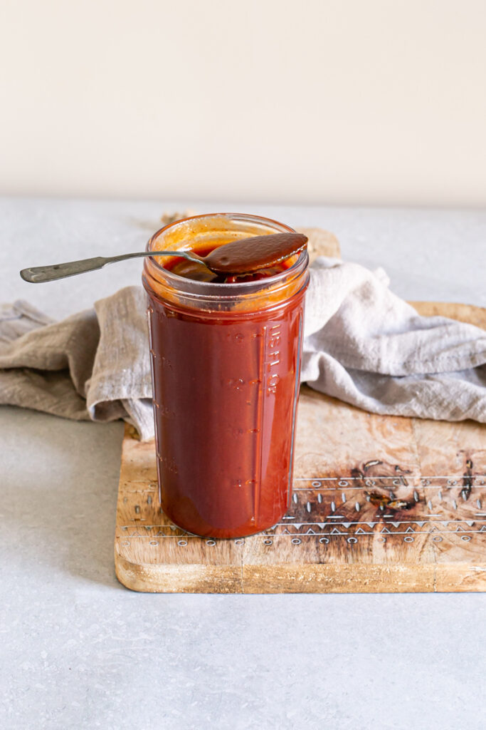 a jar of Gluten-free Vegan Pumpkin BBQ Sauce on a cutting board with a spoon resting on the opening of the jar with bbq sauce on it