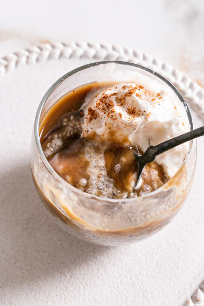 3/4 image of a glass of caramel eggnog chia pudding topped with vegan caramel, maple syrup and whipped cream with a spoonful taken out of it and the spoon in the glass
