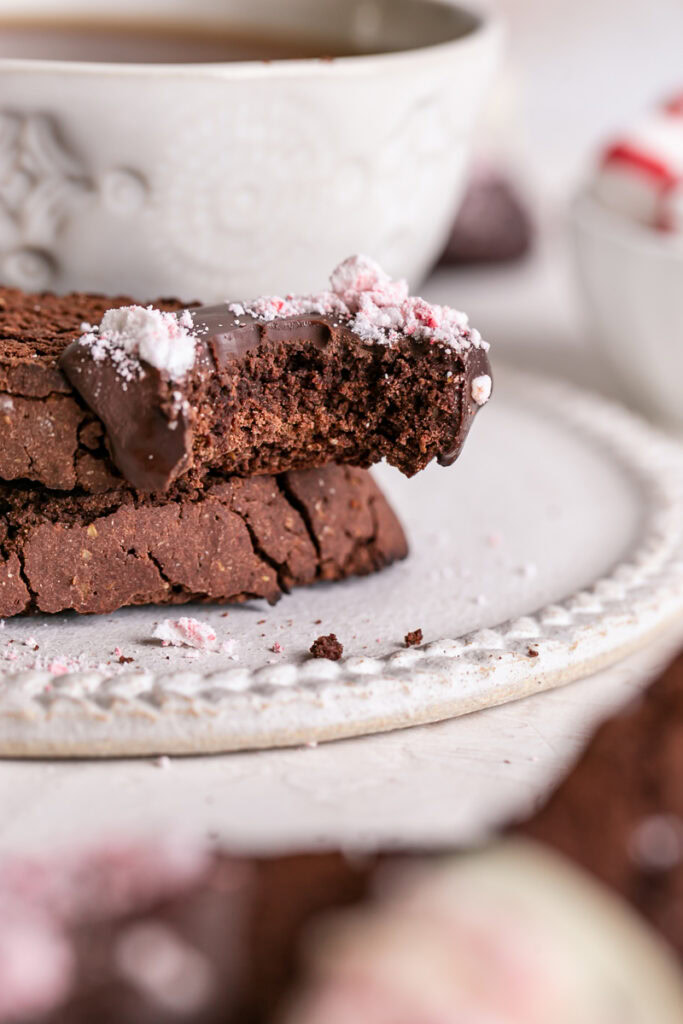 a close up vertical shot of Gluten-Free Chocolate Peppermint Biscotti with a bite taken out of it and a cup of coffee in the background