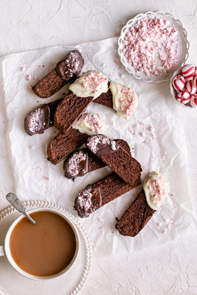 Gluten-Free Chocolate Peppermint Biscotti laid out on a piece of parchment paper with crush peppermints on a plate, whole peppermints in a bowl and a cup of coffee 