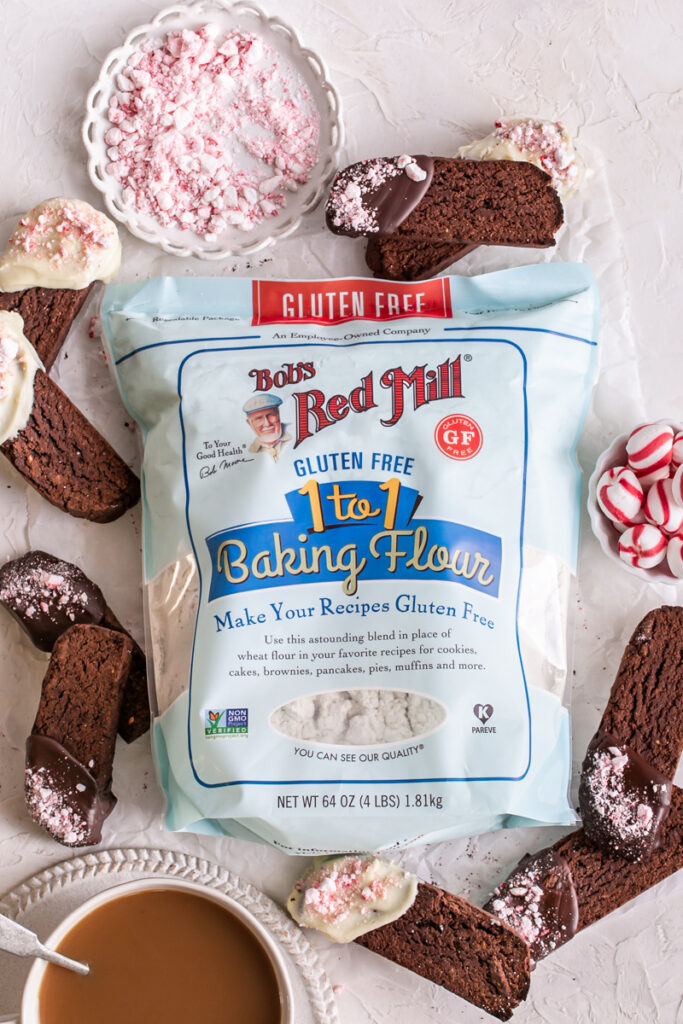 bob's red mill gluten-free 1-to-1 baking flour surrounded by Gluten-Free Chocolate Peppermint Biscotti, a plate of crushed peppermints and a cup of coffee