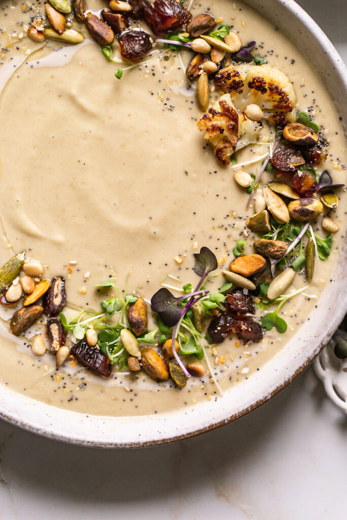 a close up shot of a bowl of roasted cauliflower soup garnished with micro greens roasted nuts, dates and everything bagel seasoning