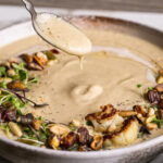 close up action shot of a bowl of roasted cauliflower soup with a spoon dripping some off of it