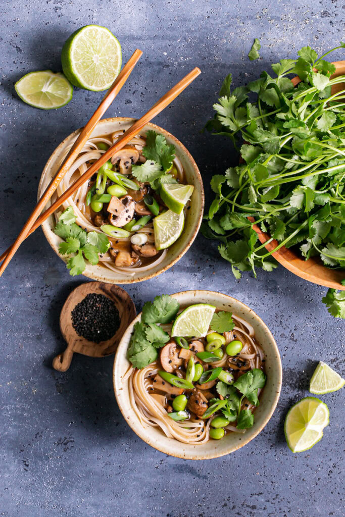 two bowls of mushroom pho garnished with fresh cilantro, green onion, black sesame seeds, lime wedges and a bowl of fresh cilantro next to it