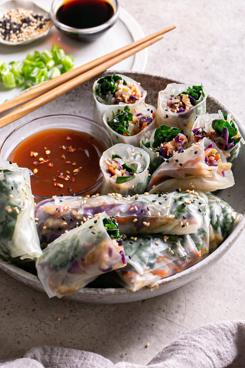 3/4 view of a bowl of sesame kale spring rolls, some cut in half to reveal the filling, served with a bowl of sweet and sour sauce