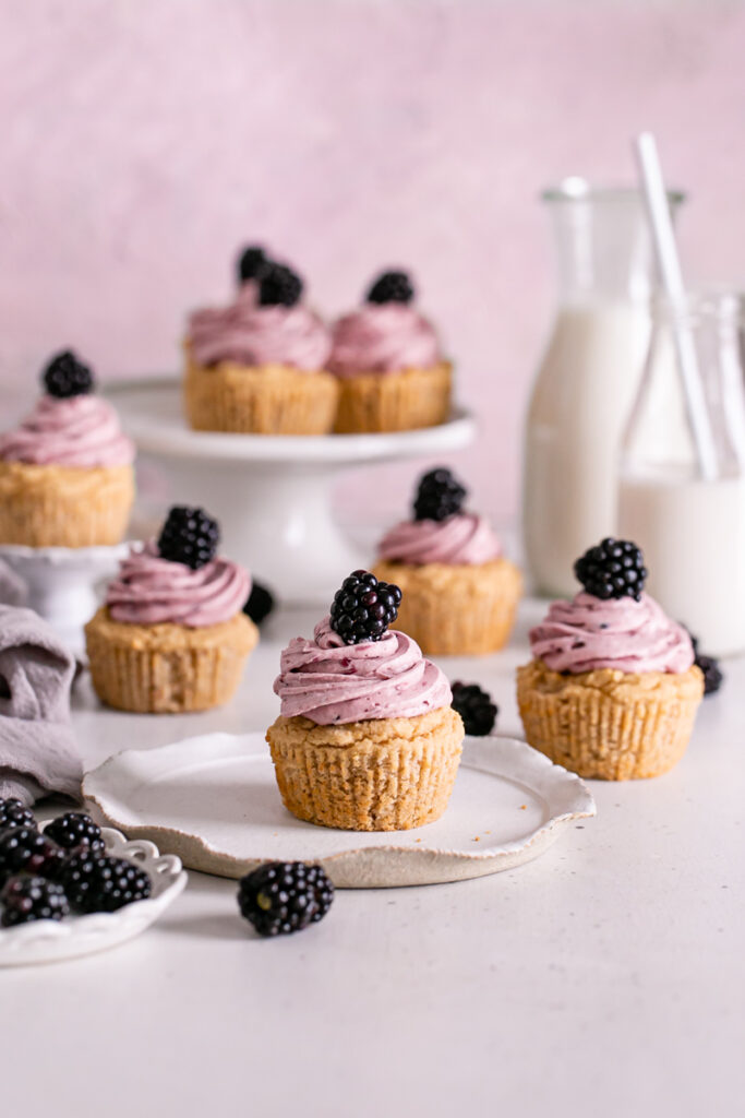 blackberry frosted cupcakes topped with a fresh blackberry, a plate of fresh blackberries in front and two jars of almond milk in the background 