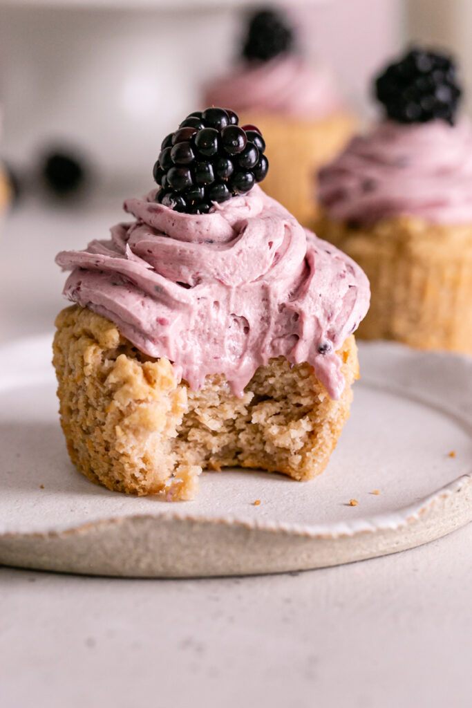 a blackberry frosted vanilla cupcake on a plate with a blackberry on top and a bite taken out of it. 