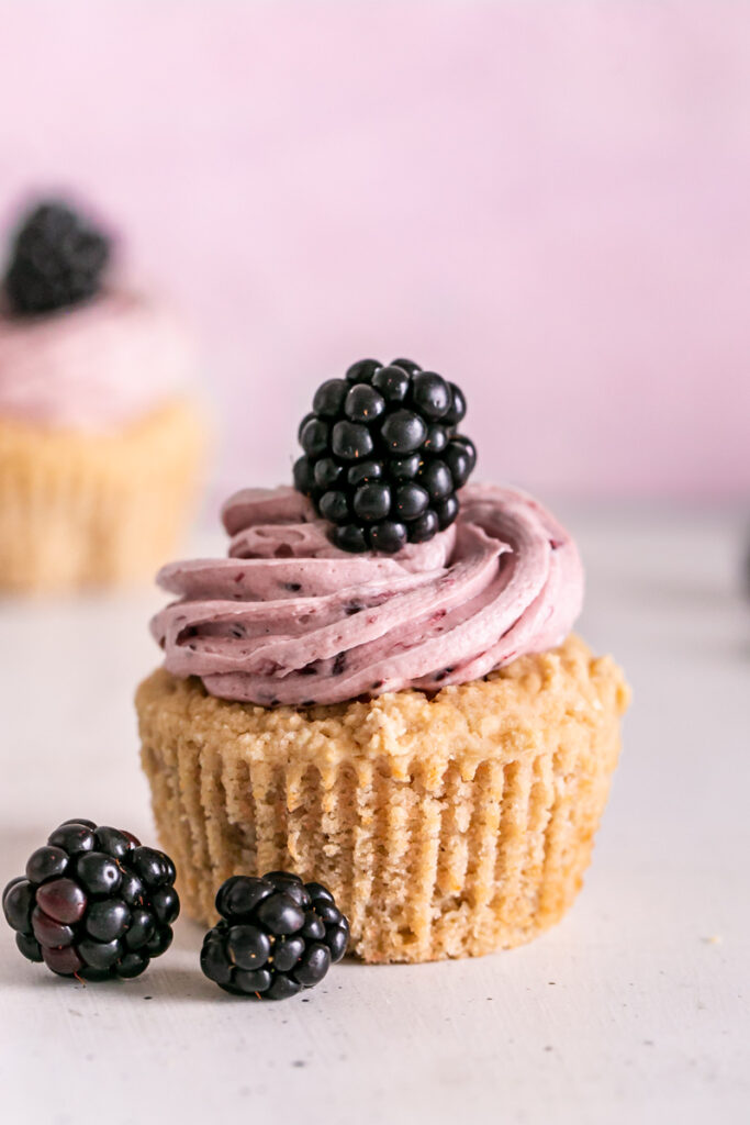 a single vanilla cupcake with blackberry buttercream frosting, a fresh blackberry on top and two blackberries lying in front of it