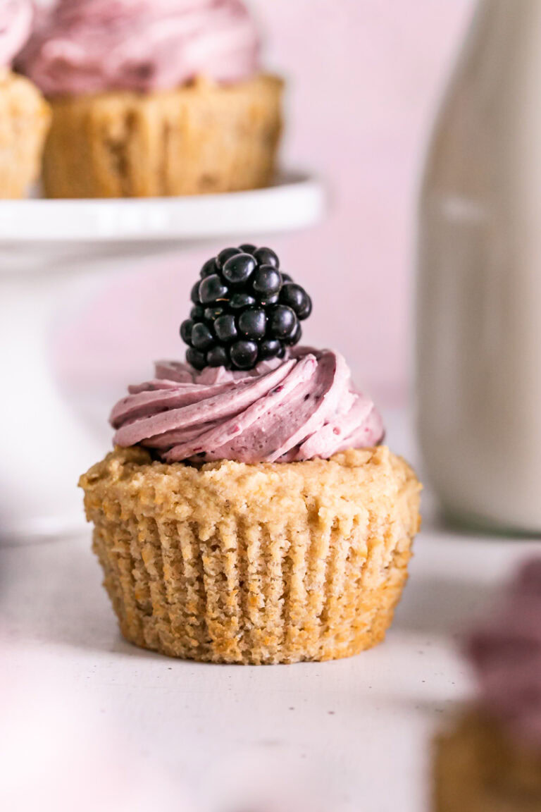 a single vanilla frosted cupcake with a blackberry on top, a jar of almond milk in the background and a cake stand of more cupcakes in the background