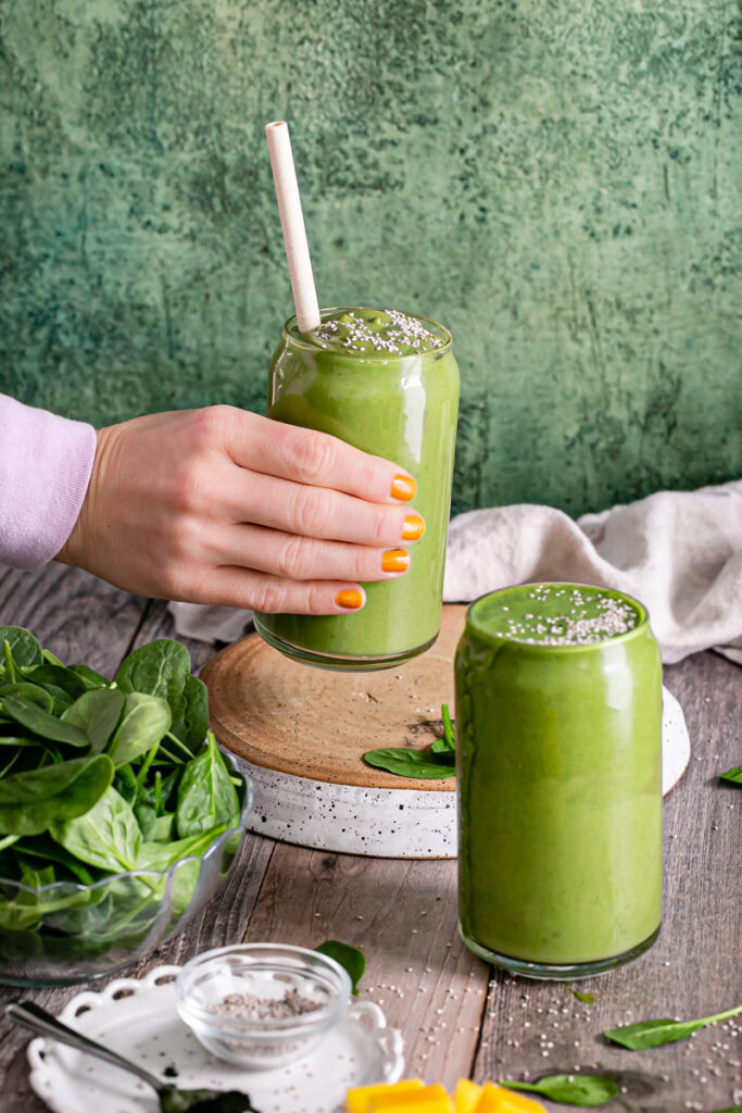hand holding up a Anti-Inflammatory Vegan Green Smoothie with a straw in it and another glass of smoothie in front. A large bowl of spinach and a linen napkin in the background