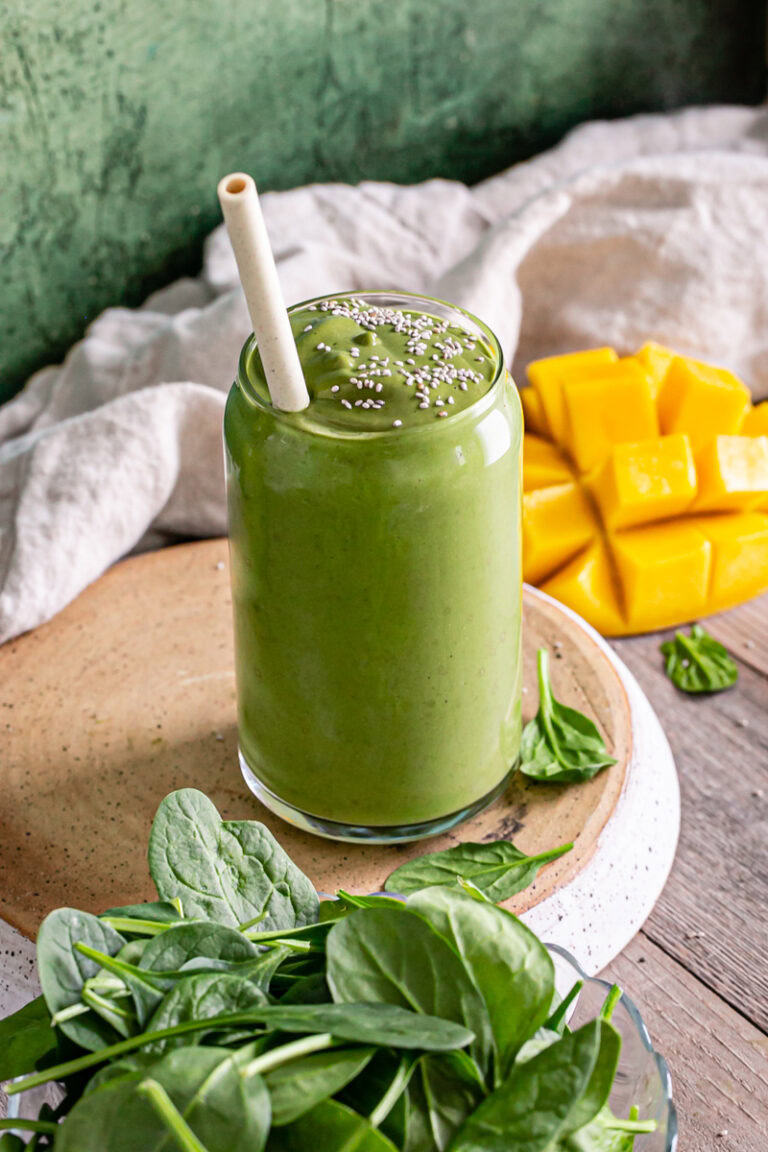 3/4 view of a single Anti-Inflammatory Vegan Green Smoothie with a bowl of spinach in front and fresh mango in the background