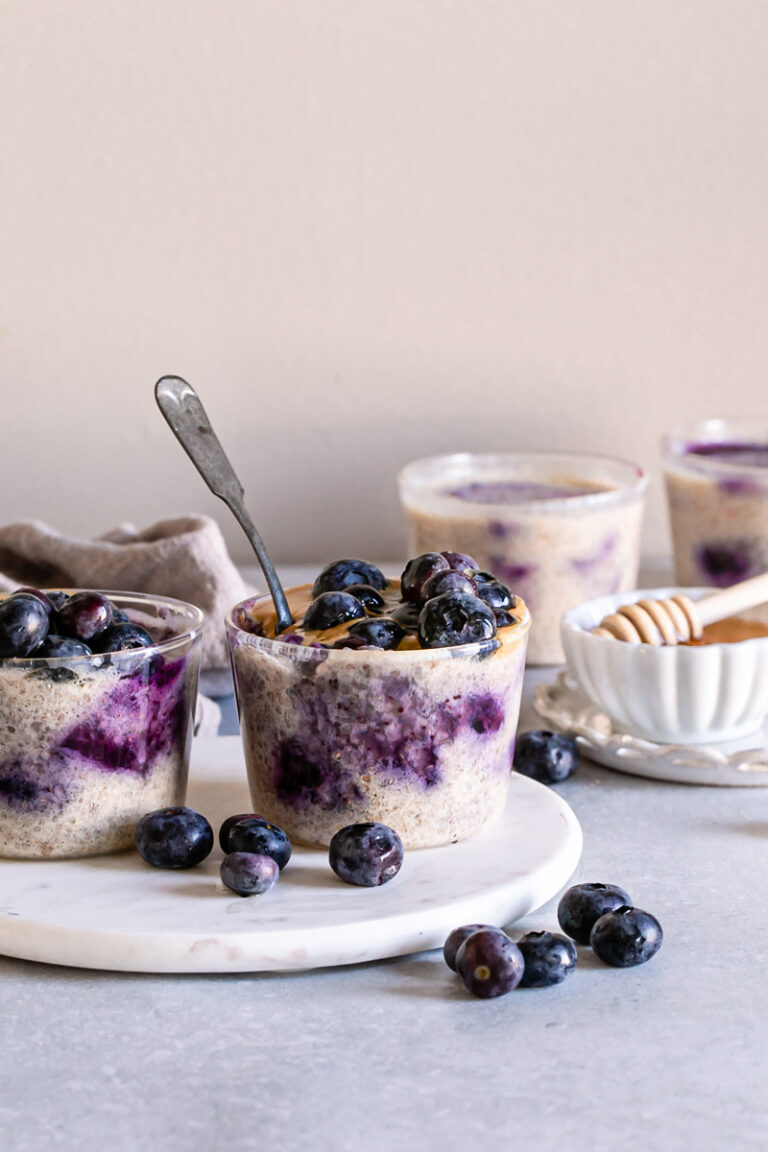 two cups of Blueberry Overnight Quinoa with fresh blueberries on top and around the glasses, a bowl of agave syrup and a honey stick behind it