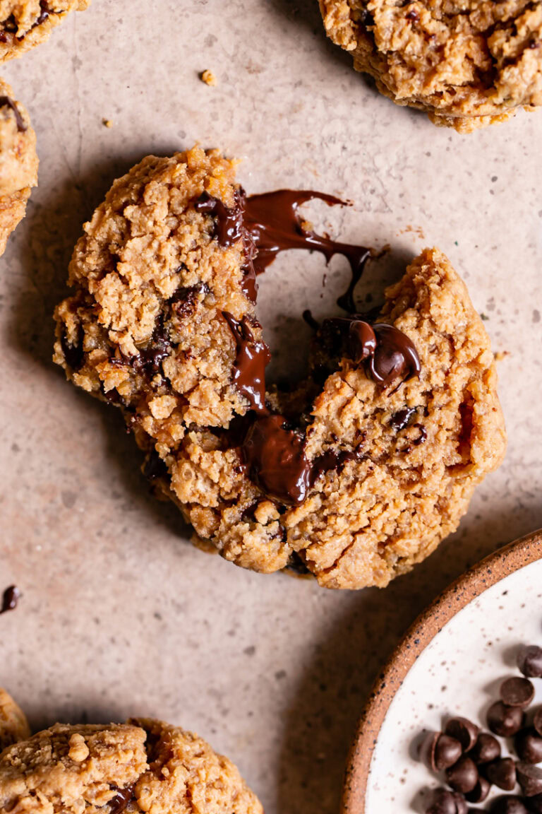 a Quinoa Chocolate Chip Cookie broken with melted chocolate oozing from the middle and a plate of chocolate chips in the corner with other cookies around it