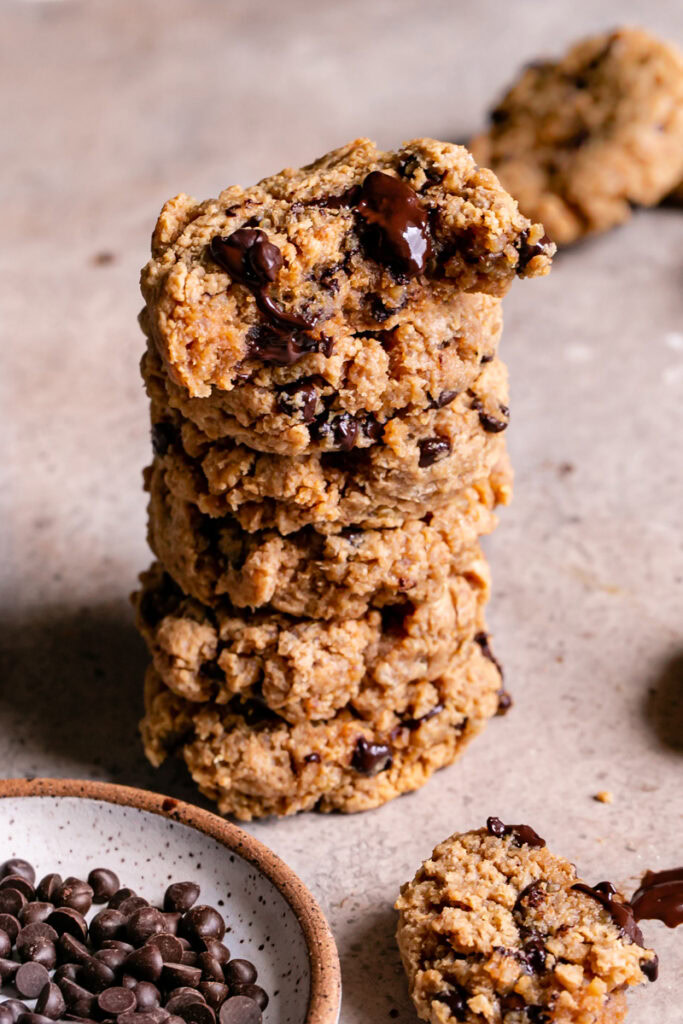 Quinoa Chocolate Chip Cookies stacked on top of one another with a plate of chocolate chips in front and the top cookie with a bite taken out of it