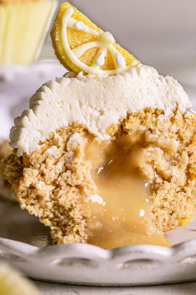 close up of a frosted grain-free lemon cupcake topped with a candy lemon wedge cut in half to reveal the lemon curd filling oozing from the middle 