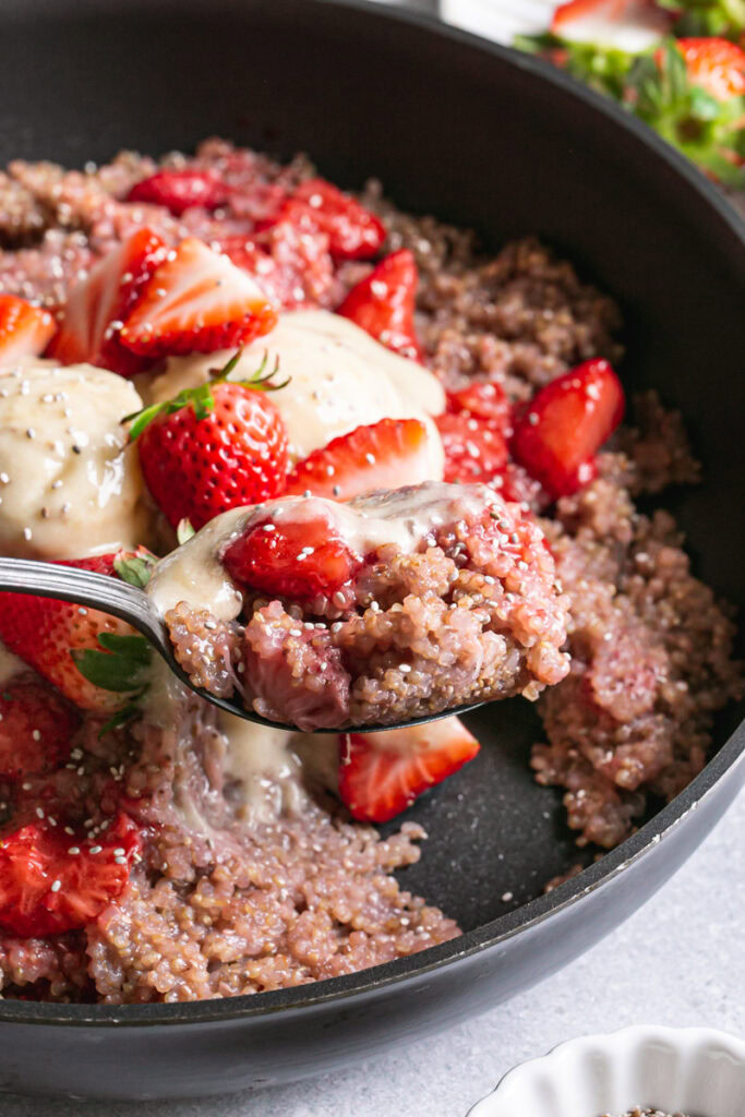a close up of a spoon spooning some strawberries and cream breakfast quinoa out of the skillet