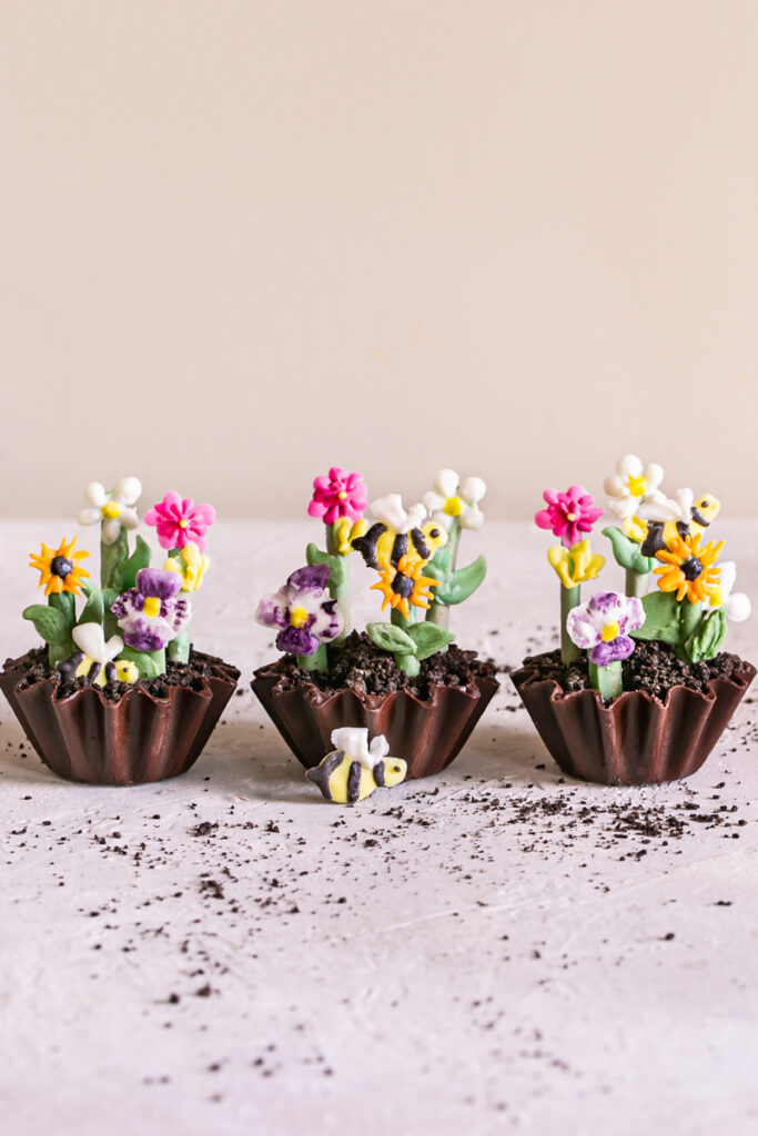 three Chocolate Avocado Pudding Dirt Cups decorated with painted candy flowers and bees with Oreo crumbs sprinkled around them