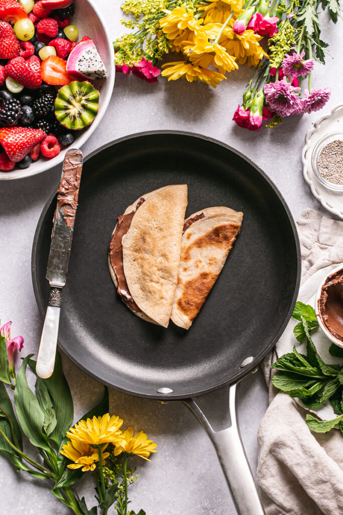 gluten-free vegan crepes folded over with chocolate hazelnut spread in the calphalon fry pan a bowl of fresh fruit above it, flowers surrounding it