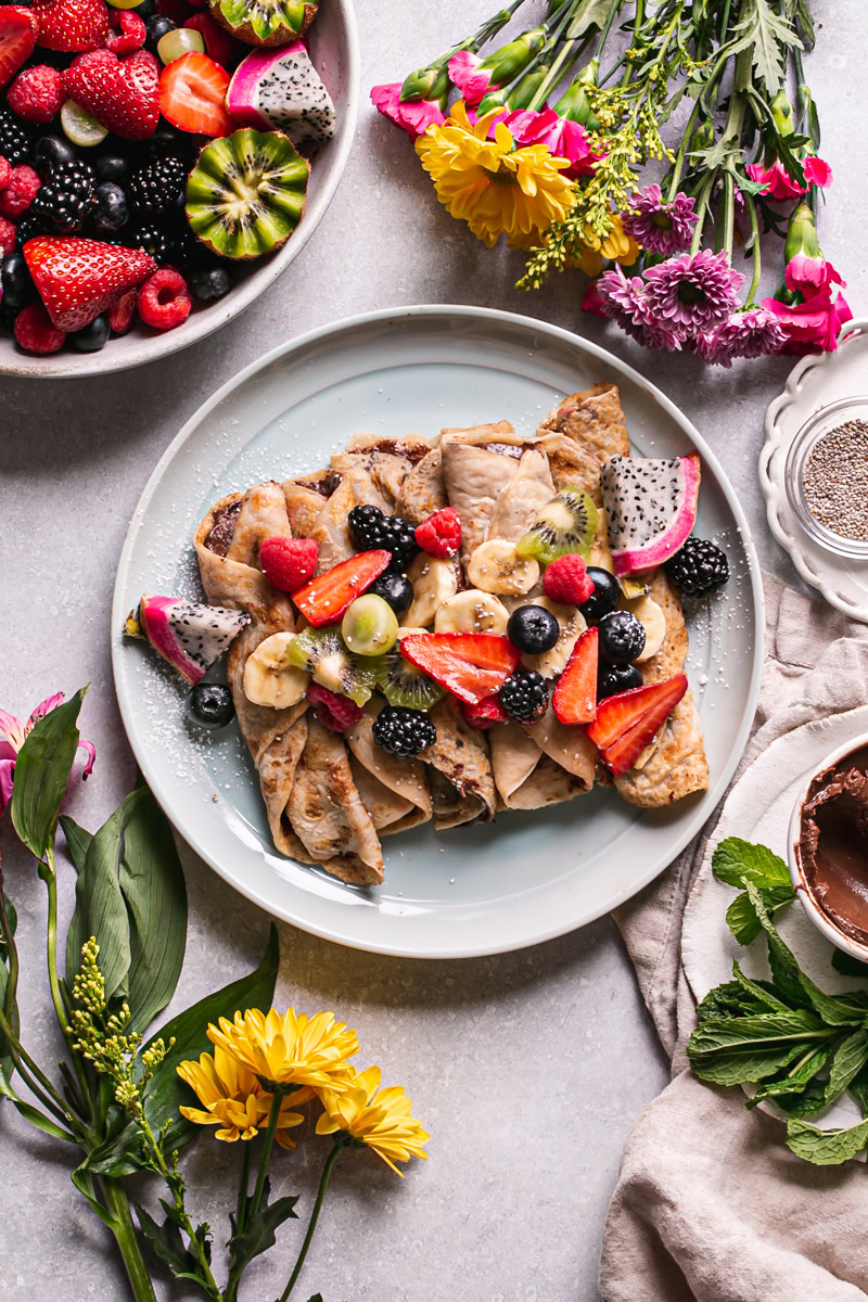 overhead shot of gluten-free vegan crepes rolled up on a plate with fresh fruit on top of them and a bowl of fresh fruit in the corner above the plate and fresh flowers surrounding the plate
