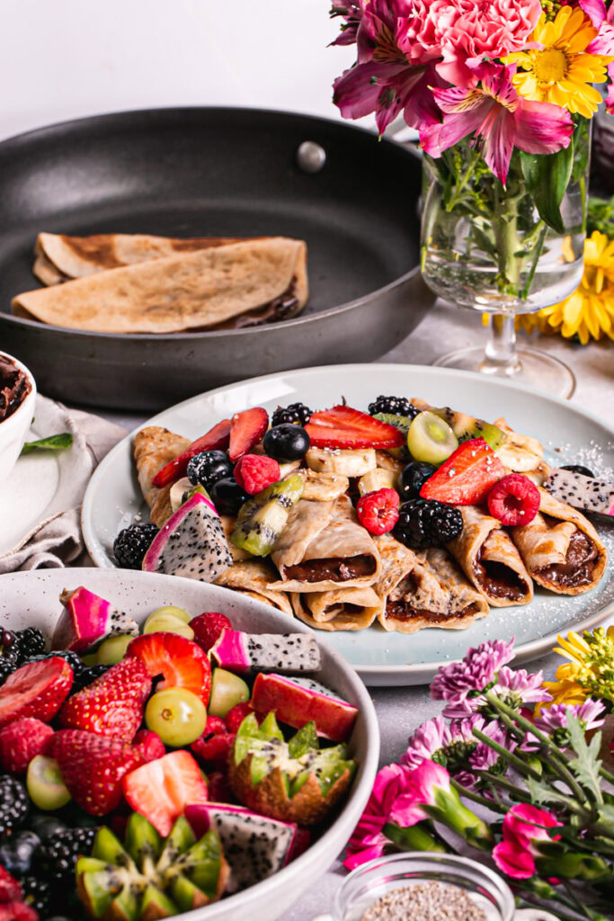 gluten-free vegan crepes rolled up on a plate with fresh fruit on top, a bowl of fresh fruit in front, the calphalon fry pan in the background with two crepes in it and flowers all around it