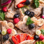 close up of chocolate hazelnut gluten-free vegan crepes folded on a wooden board topped with fresh fruit, a bowl of chocolate hazelnut spread