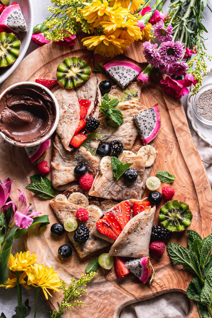 chocolate hazelnut gluten-free vegan crepes folded on a wooden board topped with fresh fruit, a bowl of chocolate hazelnut spread and fresh flowers surrounding the board