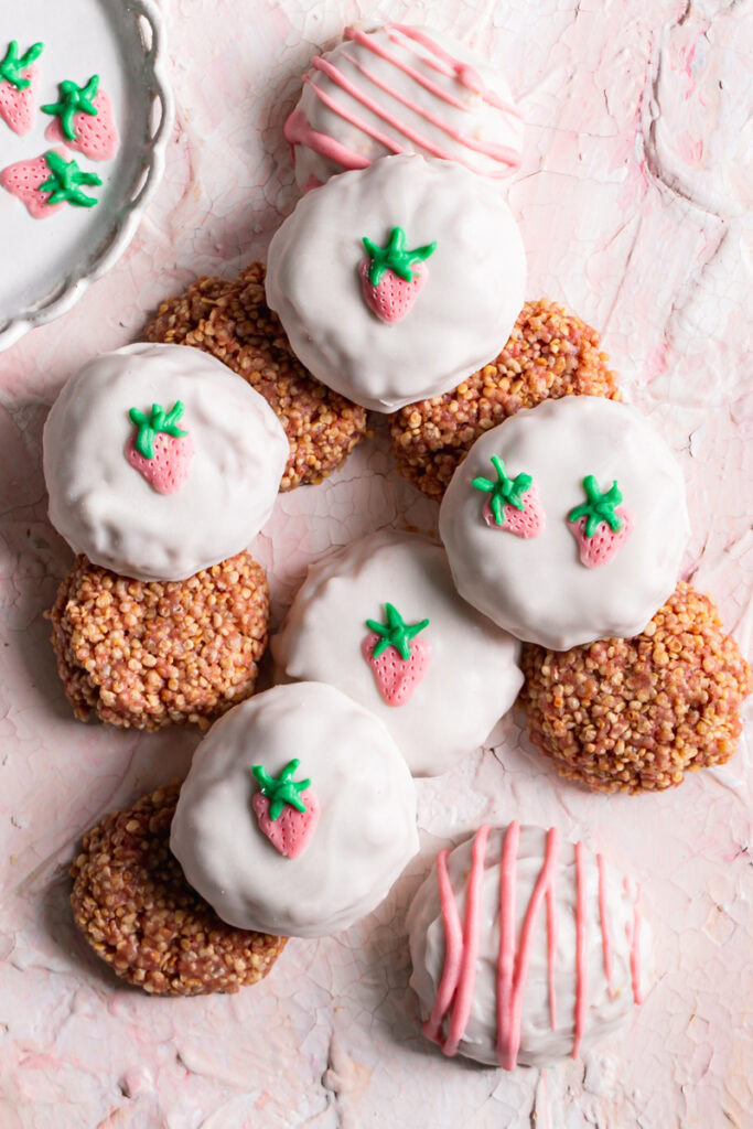 strawberries and cream qunioa cookies piled on top of one another, some covered in white chocolate decorated with a white chocolate strawberry and others not coated in white chocolate to reveal the quinoa cookie texture 