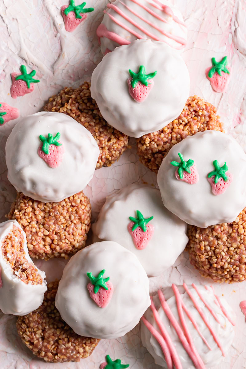 close up of strawberries and cream qunioa cookies piled on top of one another, some covered in white chocolate decorated with a white chocolate strawberry and others not coated in white chocolate to reveal the quinoa cookie texture