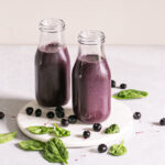 two mini milk jars filled with blueberry spinach spirulina smoothie on a round marble plate with blueberries and spinach leaves sprinkled around it