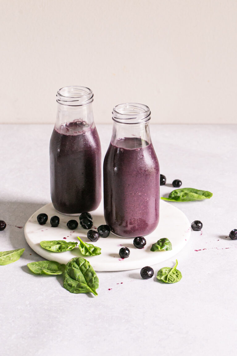 two mini milk jars filled with blueberry spinach spirulina smoothie on a round marble plate with blueberries and spinach leaves sprinkled around it