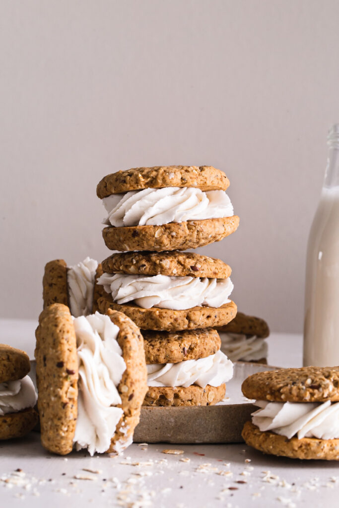 three oatmeal cookie sandwiches stacked on top of one another with another on the side and one in front with a jug of plant milk in the background