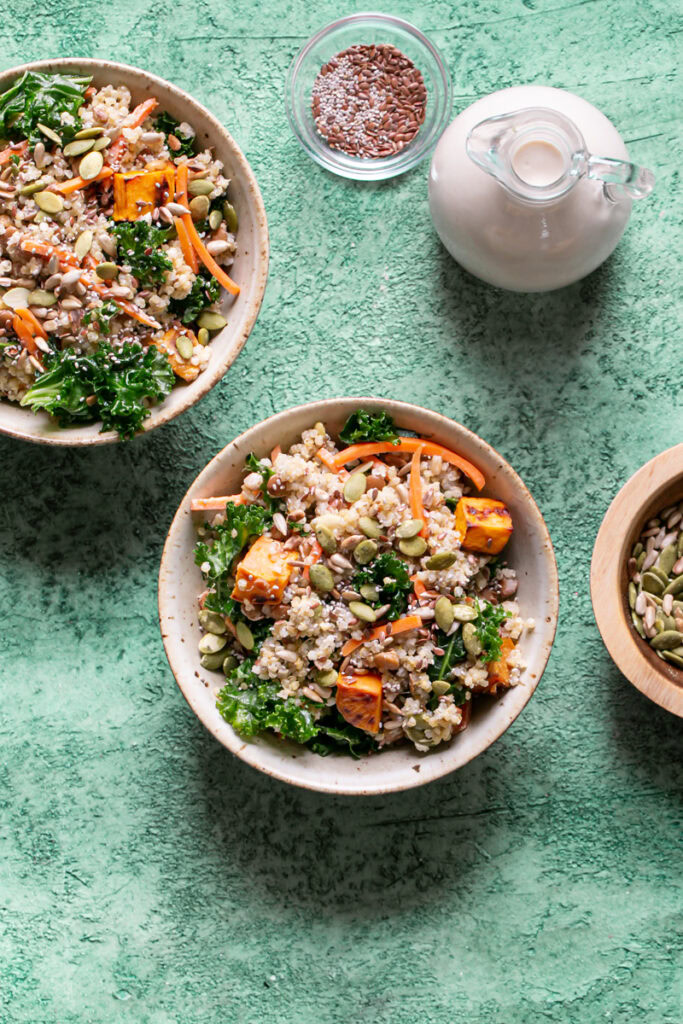 two bowls of Protein Packed Creamy Tahini Quinoa Bowls on a green backdrop with a bowl of seeds above and beside them with the tahini dressing in a bottle above the bowls