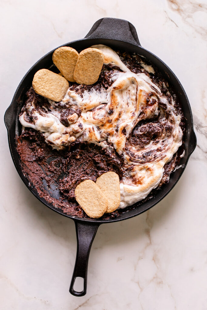 Vegan Cast Iron S'mores Dip in a Calphalon cast iron skillet with toasted vegan marshmallow fluff and gluten-free graham crackers laid inside the skillet and some of the dip already eaten