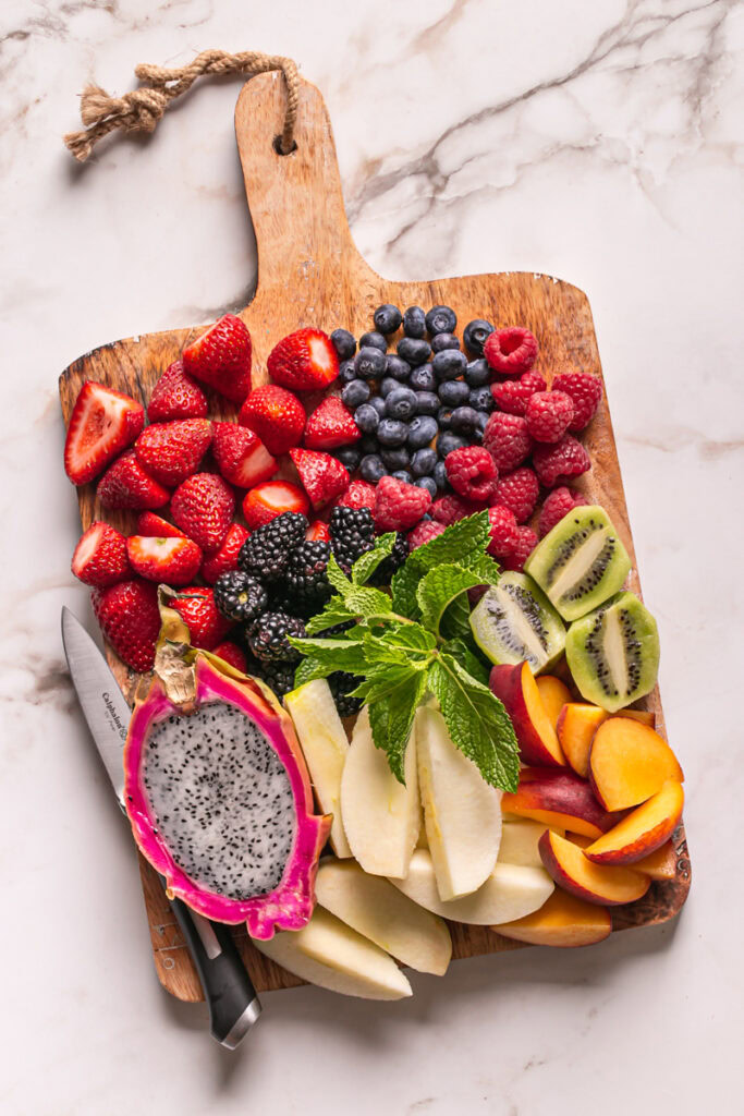 strawberries, blueberries, raspberries, blackberries, kiwi, apple, peach, dragonfruit, and fresh mint leaves on a cutting board with a calphalon knife laying beside the fruit