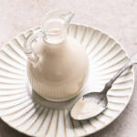 creamy tahini dressing in a glass oil bottle on a plate with a spoon with some of the dressing on it on the plate beside the bottle