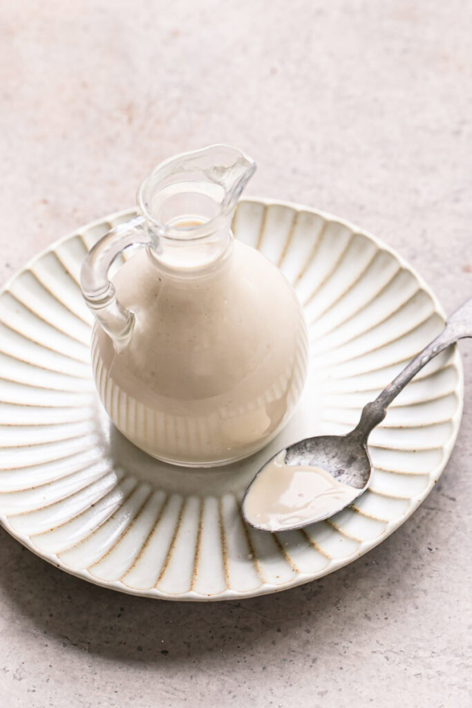 creamy tahini dressing in a glass oil bottle on a plate with a spoon with some of the dressing on it on the plate beside the bottle